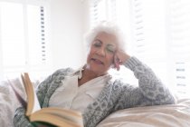 Femme âgée de race mixte assis sur le canapé livre de lecture. rester à la maison dans l'isolement pendant le confinement en quarantaine. — Photo de stock