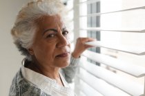 Портрет пожилой женщины смешанной расы, смотрящей в камеру у окна. оставаться дома в изоляции во время карантинной изоляции. — стоковое фото
