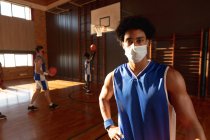 Portrait of mixed race male basketball player wearing face mask with team in background. basketball, sports training at an indoor court during coronavirus covid 19 pandemic. — Stock Photo