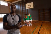 Retrato del entrenador de baloncesto afroamericano que sostiene el portapapeles con el equipo en el fondo. baloncesto, entrenamiento deportivo en una cancha cubierta. - foto de stock