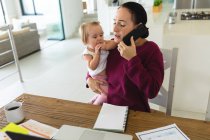 Кавказька мати тримає своє немовля, розмовляючи на смартфоні, працюючи вдома. материнство, любов і догляд дітей — стокове фото