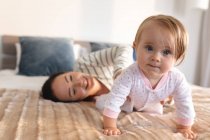 Retrato del bebé caucásico sentado en la cama mientras la madre la sostiene en la cama en casa. maternidad, amor y cuidado del bebé - foto de stock