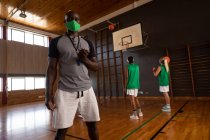 Retrato de un entrenador de baloncesto afroamericano usando máscara facial con el equipo en el fondo. baloncesto, entrenamiento deportivo en una cancha de interior durante la pandemia de coronavirus covid 19. - foto de stock
