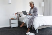 Mixed race senior woman sitting on bed using laptop. staying at home in isolation during quarantine lockdown. — Stock Photo