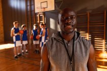 Portrait de l'entraîneur afro-américain de basket-ball masculin avec l'équipe en arrière-plan. basket-ball, entraînement sportif sur un terrain intérieur pendant. — Photo de stock