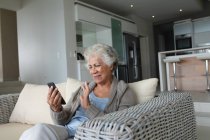 Femme âgée de race mixte assise sur un canapé faisant un appel vidéo à l'aide d'un smartphone. rester à la maison dans l'isolement pendant le confinement en quarantaine. — Photo de stock