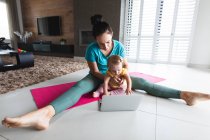 Madre caucásica sosteniendo a su bebé realizando ejercicio de estiramiento mirando a la computadora portátil en casa. maternidad, amor y cuidado del bebé - foto de stock