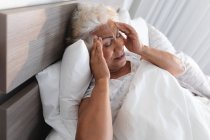 Змішана раса старша жінка лежить у ліжку, тримаючи голову в думці. перебування вдома в ізоляції під час карантину . — стокове фото