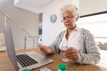 Femme âgée de race mixte assis à la table en utilisant un ordinateur portable et en prenant des médicaments. rester à la maison dans l'isolement pendant le confinement en quarantaine. — Photo de stock