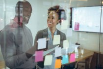Zwei unterschiedliche Geschäftskollegen lächeln und schreiben auf eine Glaswand Notizen. Arbeit in einem modernen Büro. — Stockfoto