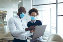 Two diverse business colleagues wearing face masks and using laptop. work at a modern office during covid 19 coronavirus pandemic. — Stock Photo