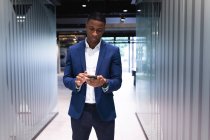 Hombre de negocios afroamericano usando un teléfono inteligente mientras está de pie en la oficina moderna. concepto de negocio y oficina - foto de stock