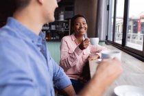 Couple mixte tenant des tasses à café souriant tout en étant assis dans un café. couple date et relation concept — Photo de stock