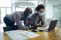 Дві різні чоловічі та жіночі бізнес-колеги носять маски для обличчя та використовують ноутбук. робота в сучасному офісі під час пандемії коронавірусу 19 . — стокове фото