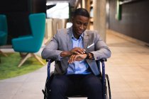 Ein behinderter afrikanisch-amerikanischer Geschäftsmann, der in einem modernen Büro im Rollstuhl sitzt und eine Smartwatch benutzt. Geschäfts- und Bürokonzept — Stockfoto