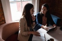 Две кавказские женщины сидят за столом с кофе, за планшетом, разговаривают. небольшой независимый кафе-бизнес. — стоковое фото