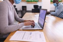 Two diverse male and female business colleagues wearing face masks and using laptops. work at a modern office during covid 19 coronavirus pandemic. — Stock Photo