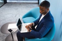 Hombre de negocios afroamericano con portátil usando smartwatch mientras está sentado en una silla en la oficina moderna. concepto de negocio y oficina - foto de stock