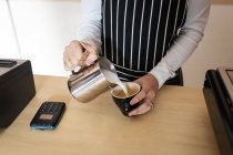 Midsection of female business owner wearing apron, pouring froth milk to coffee. small independent cafe business. — Stock Photo