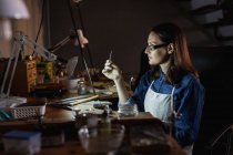 Caucasian female jeweller wearing apron, and glasses, sitting at desk, making jewelry in workshop. independent handmade craft business. — Stock Photo