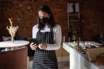 Imprenditrice caucasica che indossa maschera facciale e grembiule, utilizzando tablet. piccola caffetteria indipendente durante coronavirus covid 19 pandemia. — Foto stock