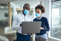 Два різних бізнес-колеги носять маски для обличчя та використовують ноутбук. робота в сучасному офісі під час пандемії коронавірусу 19 . — стокове фото