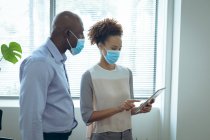 Zwei unterschiedliche Geschäftskollegen tragen Gesichtsmasken und nutzen Tablets. Arbeit in einem modernen Büro während der Covid 19 Coronavirus-Pandemie. — Stockfoto