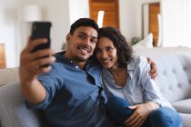 Счастливая латиноамериканская пара, сидящая на диване в гостиной, делая селфи и улыбаясь. проводить время вместе дома. — стоковое фото