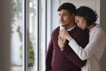 Thoughtful hispanic couple standing at window looking out and embracing. spending free time together at home. — Stock Photo