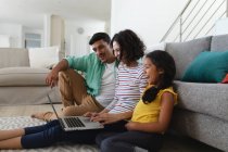 Sonriendo madre hispana, padre e hija sentados en el piso de la sala de estar usando un portátil juntos. familia pasar tiempo juntos en casa. - foto de stock