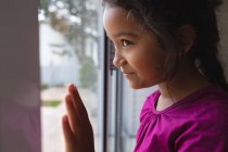Menina hispânica feliz de pé na janela com a mão no vidro, olhando para fora e sorrindo. tempo livre em casa. — Fotografia de Stock