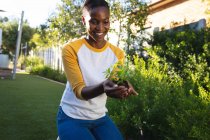 Sorridente donna afro-americana che fa giardinaggio, inginocchiata con piantina in mano a coppa nel giardino soleggiato. trascorrere del tempo libero a casa. — Foto stock