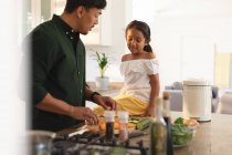 Feliz hija hispana y padre preparando verduras en la cocina, hija sentada en el mostrador. en casa en aislamiento durante el bloqueo de cuarentena. - foto de stock