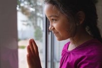 Glückliches hispanisches Mädchen, das mit der Hand auf Glas am Fenster steht, hinausblickt und lächelt. Freizeit zu Hause. — Stockfoto