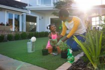 Feliz madre afroamericana y su hija de rodillas que tienden a las plantas en macetas en el jardín soleado. familia pasar tiempo juntos en casa. - foto de stock