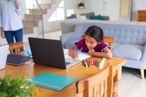 Happy hispanic girl sitting at kitchen table doing school work using laptop. happy family at home together. — Stock Photo