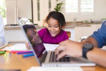 Smiling hispanic daughter sitting in kitchen doing schoolwork with father using laptop in foreground. family spending time together at home. — Stock Photo