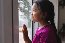 Menina hispânica feliz de pé na janela com a mão no vidro, olhando para fora e sorrindo. tempo livre em casa. — Fotografia de Stock