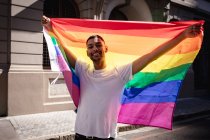 Retrato de homem gay de raça mista sorridente em pé na rua ensolarada segurando bandeira do arco-íris. igualdade de direitos e justiça manifestante em marcha de manifestação. — Fotografia de Stock