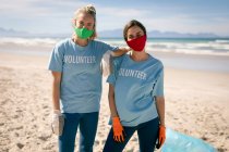 Дві різноманітні жінки у волонтерських футболках та масках для обличчя збирають сміття з пляжу. Волонтери з охорони навколишнього середовища, очищення пляжу під час пандемії коронавірусу 19 . — стокове фото