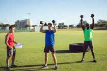 Deux hommes en forme et un entraîneur divers faisant de l'exercice à l'extérieur, soulevant des haltères. mode de vie sain et actif, entraînement croisé pour la forme physique. — Photo de stock