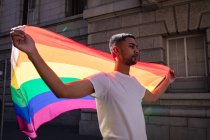 Ritratto di gay sorridente di razza mista in piedi in strada soleggiata con la bandiera arcobaleno in mano. manifestante per la parità di diritti e la giustizia in marcia dimostrativa. — Foto stock