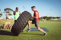 Diverse fit man and trainer exercising outdoors, encouraging and lifting heavy tyre. healthy active lifestyle, cross training for fitness. — Stock Photo