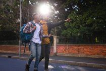 Dos felices amigos varones de raza mixta llevando mochilas caminando por la calle de la ciudad, uno señalando. vacaciones de mochilero, escapada a la ciudad. - foto de stock