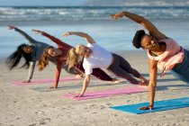 Grupo de diversas amigas practicando yoga apoyadas en un brazo que se estira en la playa. estilo de vida activo saludable, fitness al aire libre y bienestar. - foto de stock