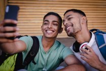 Dos amigos varones de raza mixta felices con mochilas sentadas en la calle de la ciudad tomando selfie y sonriendo. vacaciones de mochilero, escapada a la ciudad. - foto de stock
