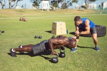 Hombre musculoso afroamericano haciendo ejercicio al aire libre con campanas de tetera e instructor de fitness. estilo de vida activo saludable, entrenamiento cruzado para fitness. - foto de stock