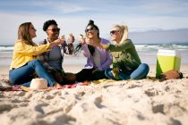 Grupo feliz de amigas divertidas se divertindo, sentadas na praia segurando a comida rindo. férias, liberdade e lazer ao ar livre. — Fotografia de Stock