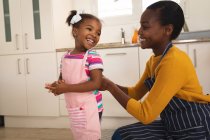 Sonriendo afroamericana madre e hija divirtiéndose en la cocina, poniéndose delantales para hornear. familia pasar tiempo juntos en casa. - foto de stock