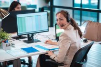 Retrato de empresária caucasiana vestindo fone de ouvido e sentada à mesa sorrindo. trabalho em um escritório moderno. — Fotografia de Stock